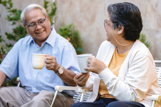Elderly couple sits in white garden chairs. They hold white tea cups, smiling at each other. Potted green plants line the stone wall. Their contented gaze shows deep companionship