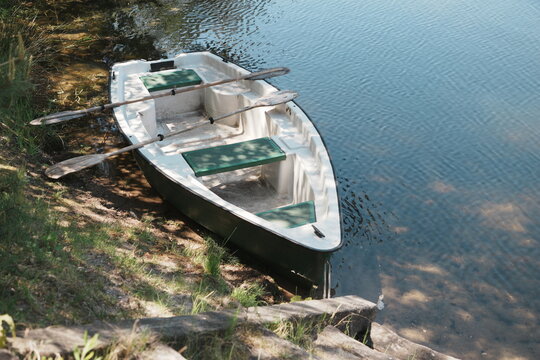 Empty White and Green Rowboat with Wooden Oars Resting on Grassy Lake Shore Ready for Peaceful Summer Recreation and Adventure