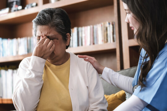 Elderly woman rubs temples in pain. Her shoulders tense with discomfort. Young caregiver places a comforting hand on her shoulder. Bookshelves line the warm home background