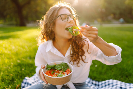 A woman sits on the grass in a sunny park, eating a fresh salad. A young woman enjoys lunch outdoors. Concepts of food, relaxation, and breaks.
