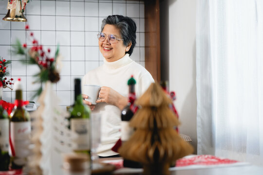 Happy elderly retired grandmother drinking tea christmas season, eyeglasses smiling winter holiday home party, lifestyle healthy aging asian grandmother kitchen environment.