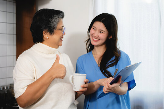 Senior woman smiles giving a thumbs-up caregiver in blue scrubs holds clipboard. They share a warm kitchen moment. Both look happy and engaged. This scene fits healthcare or senior care contexts