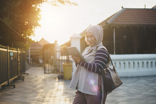 Smiling Asian Muslim Woman Standing Outdoors Using Digital Tablet During Warm Sunset Golden Hour