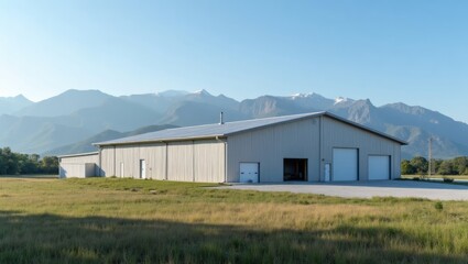 Fototapeta premium Large industrial metal building with a sloped roof situated in a rural landscape, surrounded by mountains and clear blue sky in the background