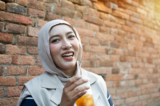 Beautiful Asian Muslim Woman Laughing Happily While Holding Iced Orange Drink Against Red Brick Wall