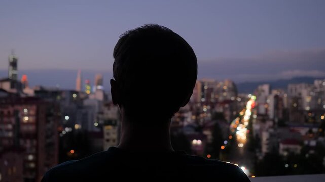 Back view: man is standing on the balcony and looking at the evening sky over the city - close up. Lonely, urban, dramatic and freedom concept
