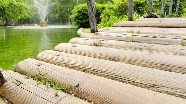 A wooden log bridge extends along a green park pond. In the background, a fountain sprays water into the air surrounded by lush trees. The daytime scene feels peaceful and calm.