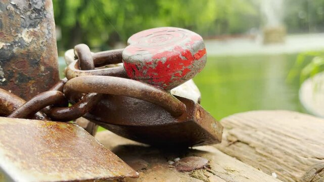 A red heart padlock and rusty locks are chained to a post. They rest on a wooden dock by a green park pond.