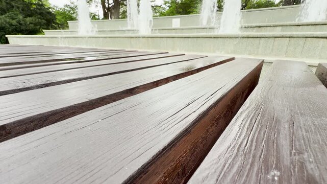 Wet wooden planks stretch out toward a multi-tiered public fountain. White water jets spray upward in a blurry green city park. The peaceful scene feels calm and refreshing.