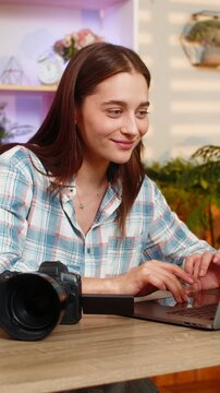 Young woman on home table edits photos on laptop near camera for creative media work. Adult lady on home table retouches images and carefully checks video clips for editing practice in living room.