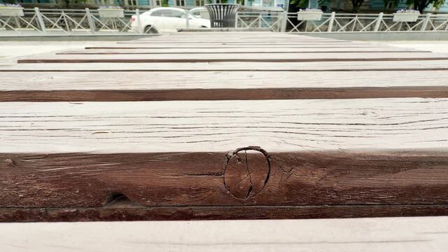 Weathered wooden planks of a park bench stretch across the frame. The background shows a blurry city street with white cars passing by. The scene is set on a quiet public plaza.