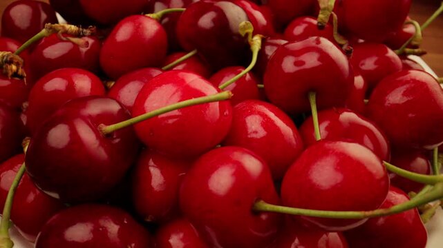 Slow panning shot closely over a pile of wet glossy ripe red sweet cherries glistening with water droplets as more fresh cherries fall onto the stack against dark background