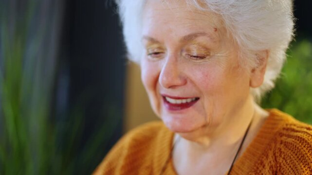 An older person sits at a table and uses a smartphone. They focus on the screen while holding the phone in their hands. The setting is simple and warm.