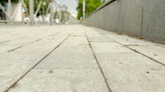 Cars scurry along the busy city road behind a metal fence. A paved sidewalk stretches forward into the urban scene.