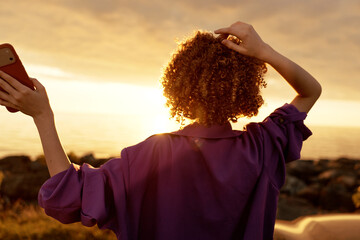 Woman with curly hair stands on a beach at sunset, taking a selfie with her phone, wearing a loose purple shirt, embodying casual lifestyle and relaxed vacation mood © SHOTPRIME STUDIO