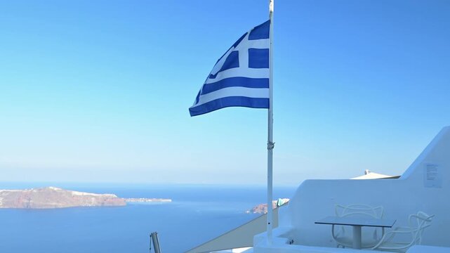 Greek flag waving over whitewashed Santorini terrace with Aegean Sea and caldera cliffs under clear summer sky.