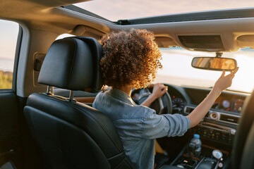 Fototapeta premium Young woman with curly hair drives a car, viewed from the back seat, sunlight streaming through the windshield, capturing a candid moment of daily commuting and focus.