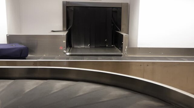 Luggage carousel at airport displays various suitcases arriving from baggage claim, showcasing a blue suitcase followed by a green bag and a purple suitcase in sequence