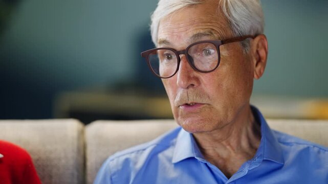 A senior man sits on a sofa in a living room. He is reading something closely while wearing glasses. The room has a bright atmosphere with light furniture.