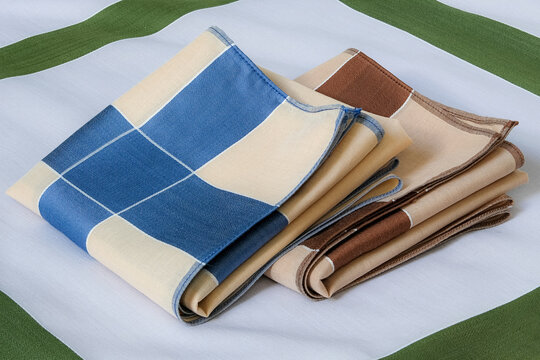 A blue-and-beige and a brown-and-beige folded handkerchiefs neatly arranged together on top of a green-bordered gray cotton handkerchief.