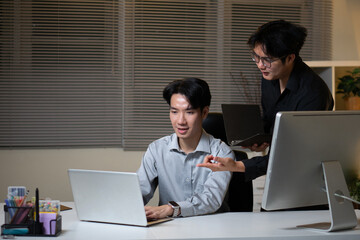 Fototapeta premium Two young men are working together at a desk, pointing at documents with pens and talking to their colleague.