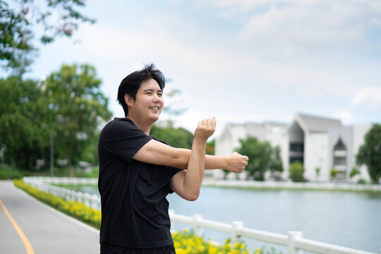 A muscular young man wearing a black T-shirt is stretching one arm across his side and crossing the other arm to warm up his shoulder muscles.