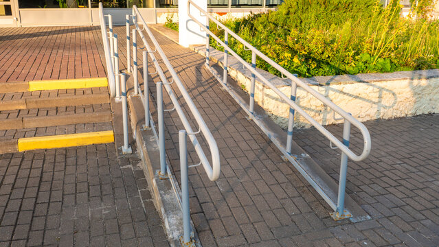 A brick-paved wheelchair accessible ramp with metal safety handrails runs parallel to outdoor concrete steps at a building entrance.