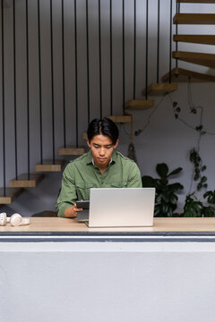 Asian man working in home workspace using silver laptop holding tablet with white headphones nearby