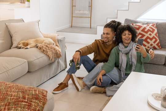 African American couple sitting on tiled floor at home, holding smartphone, dog leash, sleeping dog