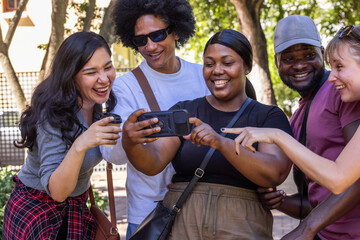 Diverse friends leaning in on paved path under trees, viewing horizontal smartphone and coffee cup © wavebreak3