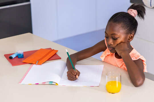 African child writing in open notebook on kitchen counter with green pencil, peach top, copy space
