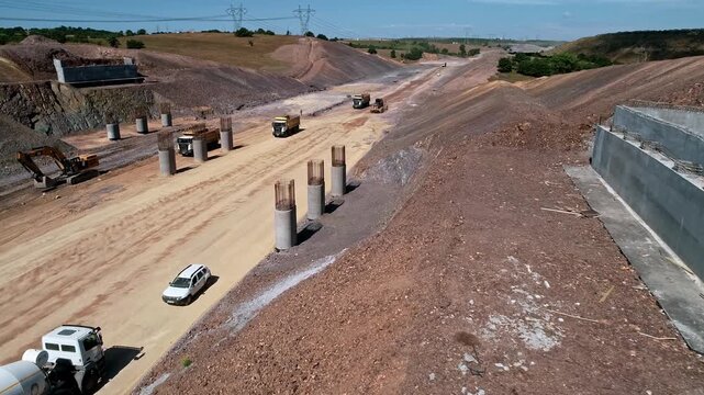 Aerial view of a road infrastructure construction site with concrete piers, retaining walls, trucks and earthworks preparing foundations for a large viaduct or elevated roadway project.