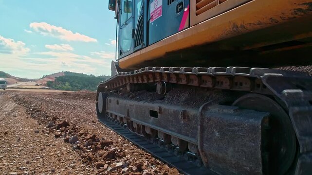Close-up view of a crawler excavator track moving on gravel ground at a construction site, showing heavy machinery undercarriage, rough terrain and earthmoving equipment detail.