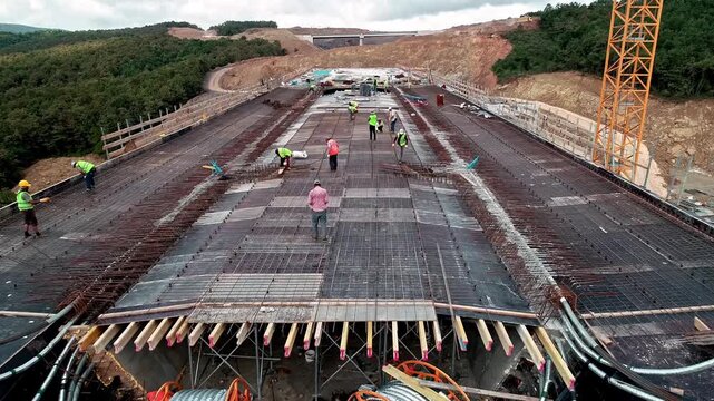 Workers installing steel reinforcement and cable systems on a large concrete deck at a road infrastructure construction site, showing structural preparation and active site work.