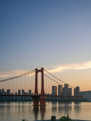 Fototapeta premium Massive red suspension bridge spanning wide calm river under soft morning sky