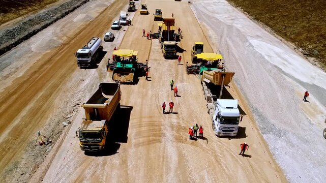 Aerial view of road paving work with dump trucks feeding material into paving machines, while workers and compactors support surface preparation at a highway construction site.