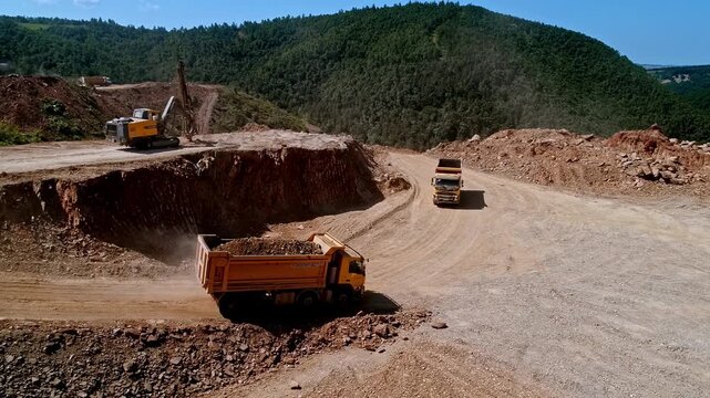 Dump trucks and drilling machinery working at a mountain excavation site, transporting soil and rock material through a dusty construction road during infrastructure development.