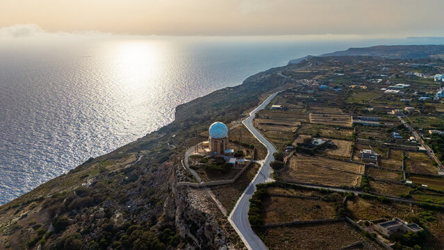 aerial drone view of dingli cliffs and radar dish dome at golden hour summer sunset with roads and green foliage below with overcast skies and Mediterranean Sea below