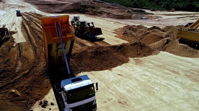 Dump truck unloading soil at a large road construction site, with bulldozer, compactor and heavy machinery preparing the ground for a highway infrastructure project.