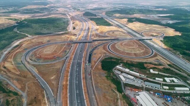 Aerial drone view of a large highway interchange with curved ramps, overpasses, traffic and surrounding forest, showing modern transportation infrastructure and road development.