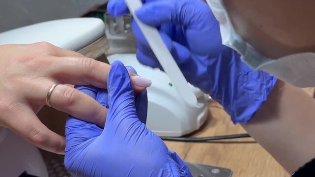 Nail technician files a client's nail with care. Blue gloves cover hands, showing professionalism. Ring gleams on client's finger, adding detail. Focused, gentle touch signals skill and comfort