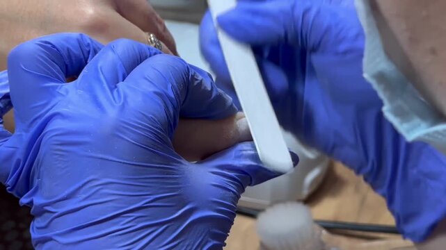 Nail technician files a client's nail with care. Blue gloves cover hands, showing professionalism. Ring gleams on client's finger, adding detail. Focused, gentle touch signals skill and comfort