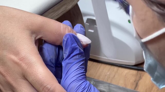 Nail technician files a client's nail with care. Blue gloves cover hands, showing professionalism. Ring gleams on client's finger, adding detail. Focused, gentle touch signals skill and comfort