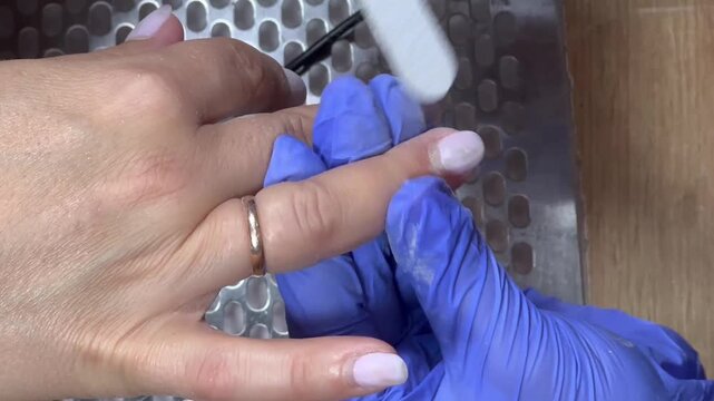 Nail technician files a client's nail with care. Blue gloves cover hands, showing professionalism. Ring gleams on client's finger, adding detail. Focused, gentle touch signals skill and comfort