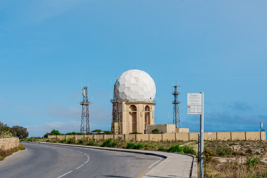 front view of dingli radar dome building with winding road and clear blue sky in summer at sunset with wall surrounding and green foliage on the dry ground at golden hour