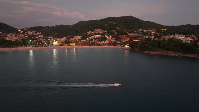A boat moves smoothly through the tranquil water, approaching a beachside town illuminated by lights, with hills in the background and a colorful parachute visible in the sky
