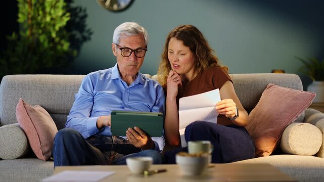 Adult daughter assisting her elderly father with online bill payments using a tablet and digital banking services. Financial management, technology support and family assistance.
