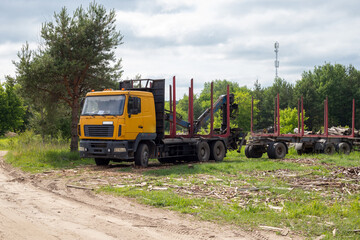 Fototapeta premium Timber truck with crane trailer at logging industry yard, yellow logging vehicle parked near forest on cloudy day