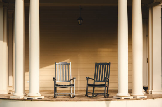 Two dark wooden rocking chairs positioned on a covered porch with elegant white columns, creating a relaxing and inviting atmosphere
