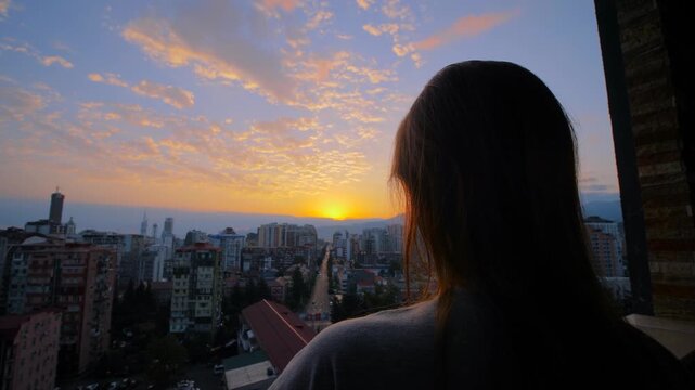 Wide angle shot: woman is standing on the balcony and looking at the dramatic sunset or sunrise sky over the city. Lonely, urban, early morning, dramatic and freedom concept